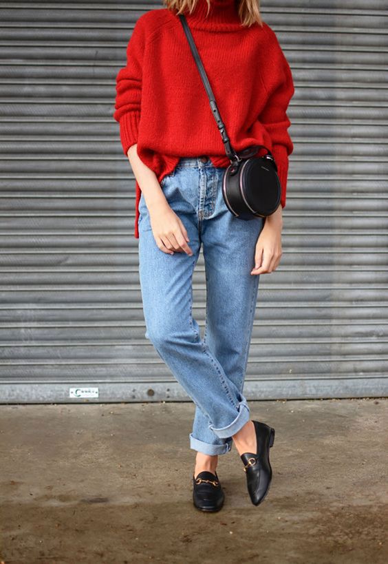 Mujer vistiendo un mom fit azúl con zapatos de piso, bolso redondo vintage y suéter rojo de cuello alto. 