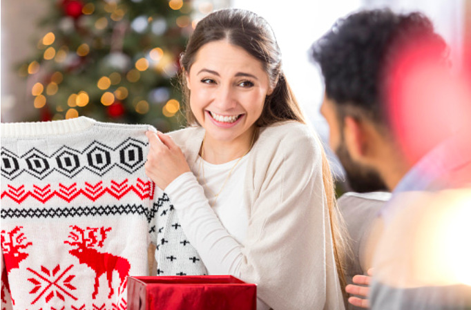 Mujer sonriente sacando de una caja de regalo un sueter blanco con adornos de navidad en color rojo y negro.